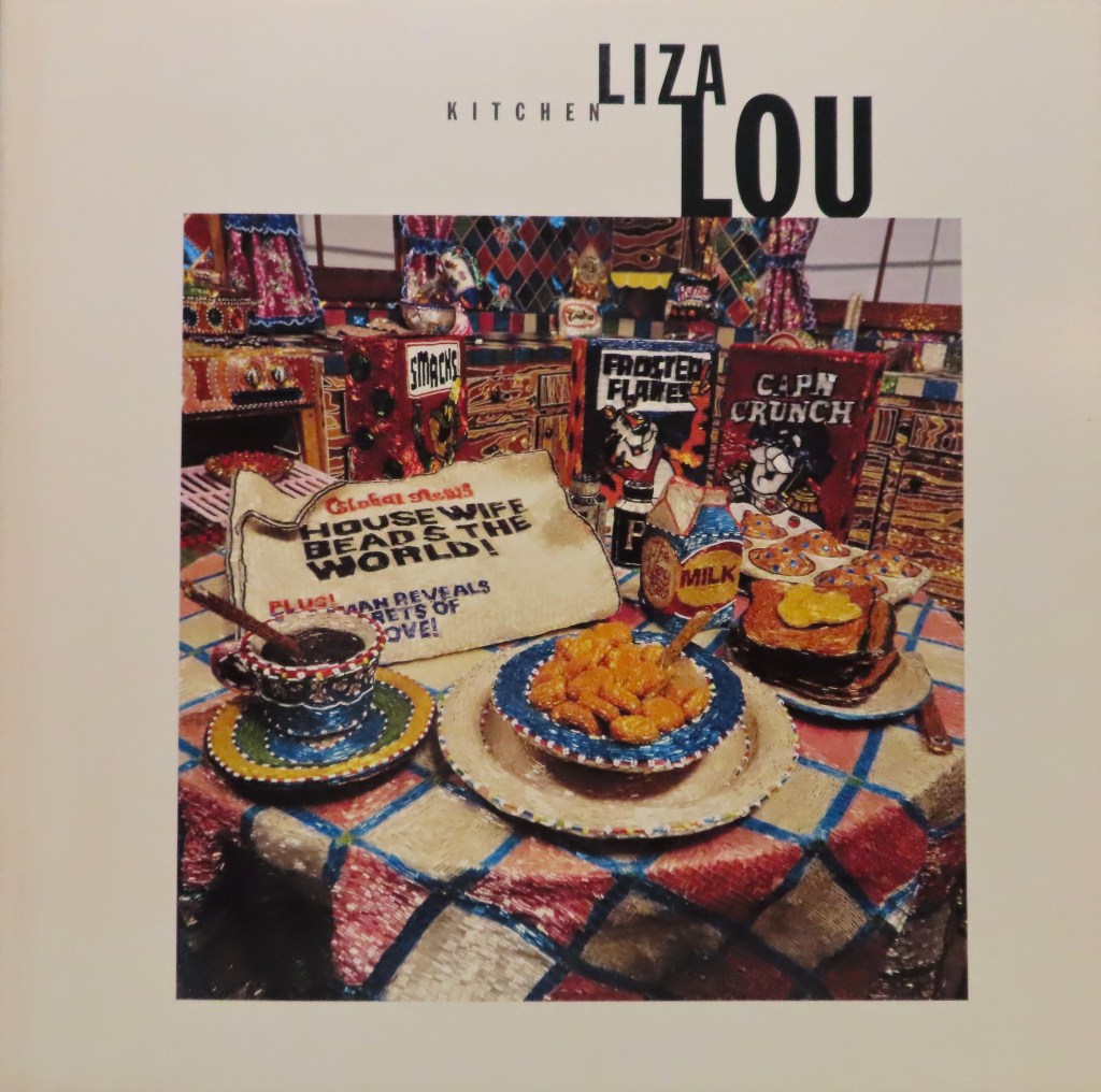 A white book cover with all capital letters that read Kitchen, Liza Lou. A brightly colored photo of one of Liza's installations a fully seqinzed kitchen with plaid round coered table, full breakfast with coffee, boxes of cereal, a newspaper that says "housewife beads the world!" Even the kitchen appliances, tiled walls, and curtains are made from sequins.