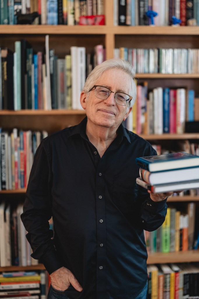 Professor Robert L. Pincus holds a stack of books. He's standing in front of a large bookcase filled with books.